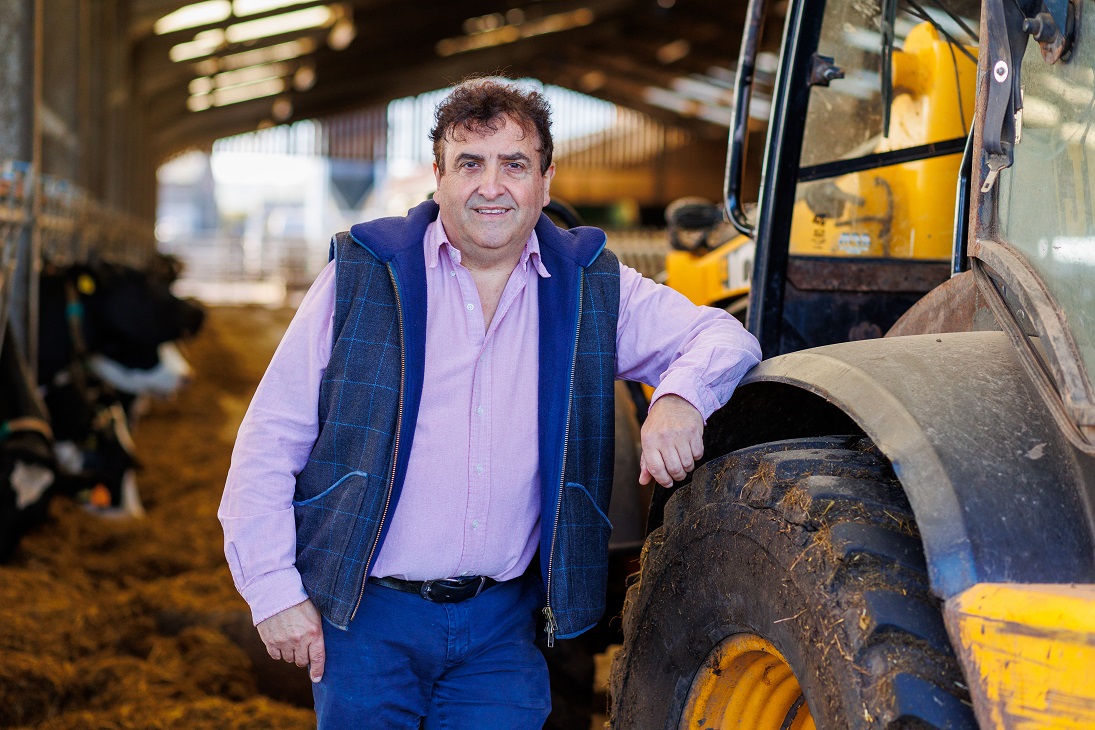 Farmer leaning on a tractor in a shed with cows eating silage in the background